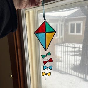 May include: A colorful stained glass kite suncatcher. The kite is diamond-shaped, with red, green, blue, and yellow panes. Below the kite are four smaller bow-tie shapes in green, red, blue, and yellow. A green cord is attached for hanging.