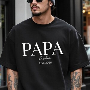 May include: Black t-shirt with the word "PAPA" in white, with "Sophia" and "EST. 2026" below. The shirt is worn by a person wearing a gray beanie and sunglasses.