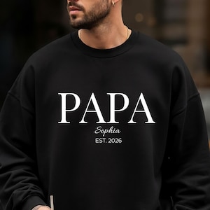 May include: Black sweatshirt with the word "PAPA" in white block letters. Below the word "PAPA" is the name "Sophia" and the year "EST. 2026" in a smaller font. The sweatshirt is a crew neck style.