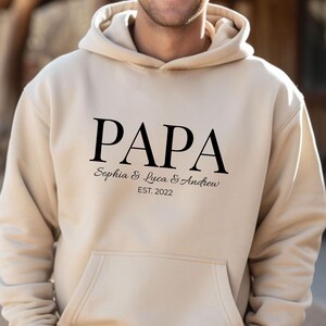 May include: A beige hoodie with a front pocket and a drawstring hood. The word "PAPA" is printed in large black letters, with "Sophia & Luca & Andrew" and "EST. 2022" below it. The person is wearing the hoodie.
