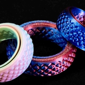 Peut inclure: Trois bagues colorées avec des surfaces texturées sont présentées sur un fond noir. Une bague est principalement arc-en-ciel pastel, une autre est rouge et bleue, et la troisième est bleue et rouge. Les bagues ont un design unique et à motifs.