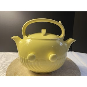 May include: A pale yellow ceramic teapot with a curved handle and a lid. The teapot has a rounded body with two circular decorative elements and a textured pattern on the lower half. The spout and handle are smooth and curved.