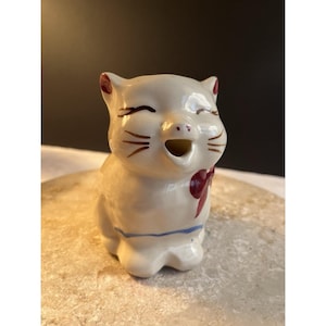 May include: A vintage ceramic cat creamer with a smiling face, red bow, and blue trim. The cat figurine is off-white with brown whiskers and ear accents. A charming addition to any kitchen or cat-themed collection.