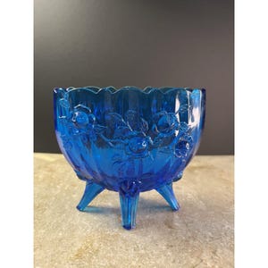 May include: A cobalt blue glass bowl featuring a rose pattern. The bowl has a scalloped edge and stands on three pointed feet. The embossed rose design is visible around the bowl's exterior. The bowl's dimensions are approximately 15 cm in diameter.