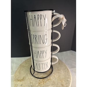 May include: A set of four white ceramic mugs stacked in a black metal stand. Each mug has a black rim and handle, with the words "HAPPY SPRING HAPPY EASTER" in black lettering. A white ribbon is tied to the top mug.