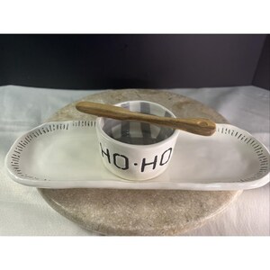 May include: A white ceramic serving set with a long, oval tray and a small bowl. The bowl has gray and white stripes and the words "HO-HO" in black. A wooden spoon rests on top.