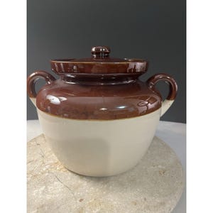 May include: A two-handled ceramic bean pot with a glossy brown glazed upper section and a smooth cream-colored lower body. The pot features a matching brown lid with a small central knob and two curved side handles for easy lifting.