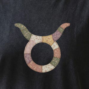 May include: Dark gray t-shirt featuring a colorful patchwork Taurus symbol. The symbol is a circle with curved horns, made of various patterned fabrics in shades of pink, green, yellow, and purple. The shirt is a casual style.