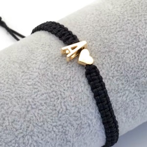 May include: Black braided bracelet with a gold-colored letter "A" charm and a heart charm. The bracelet is set against a soft gray background, highlighting the contrast between the dark cord and the bright charms.