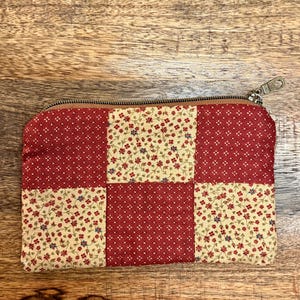 以下が含まれることがあります： ファスナー開閉の長方形パッチワークポーチ。赤とクリーム色の生地の正方形でできています。赤い正方形には小さな白いドット模様があり、クリーム色の正方形には花柄がプリントされています。