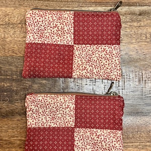 以下が含まれることがあります： 2つの小さな長方形のジッパー付きポーチ。各ポーチは、赤い花柄と赤い水玉模様の生地が交互に配置されたパッチワークデザインが特徴です。ジッパーは茶色で、ポーチは木製の表面にあります。