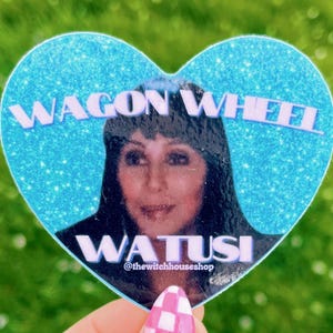 May include: A heart-shaped sticker with a glittery blue background featuring a portrait of a woman and the words "WAGON WHEEL" and "WATUSI". The sticker is held up by a hand with pink and white checkered nail polish.