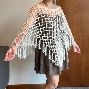May include: A cream-colored crochet poncho featuring an open net pattern and long fringe trim along the bottom edges. The garment is constructed with a wide neckline and drapes over the shoulders, showcasing a textured, hand-knitted appearance.