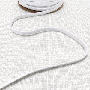 May include: A roll of white flat elastic ribbon. A length of the ribbon is unwound and lies in front of the roll. The elastic is made of a woven material and is a bright white colour. The roll is made of cardboard.
