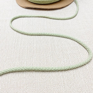 May include: A spool of light green braided cotton rope sits on a brown cardboard base. The rope is unraveled, with a length of it extending across a textured, off-white surface. The rope is a soft, muted green color.