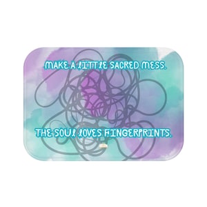 May include: Rectangular bath mat with a watercolor design in shades of purple, blue, and teal. The mat features the text "MAKE A LITTLE SACRED MESS. THE SOUL LOVES FINGERPRINTS." in a light blue font, with a tangled line design.