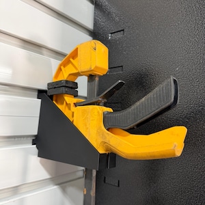 Puede incluir: Una abrazadera amarilla y negra, probablemente para carpintería o construcción, está montada sobre una superficie metálica negra. La abrazadera tiene un mango negro y una barra de metal. El fondo presenta paneles horizontales blancos.