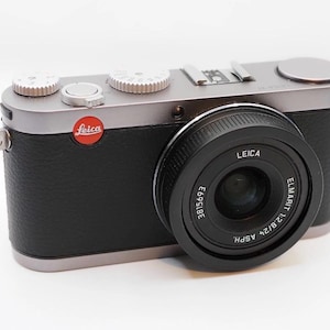 Könnte beinhalten: Silberne und schwarze Leica Digitalkamera mit schwarzem Objektiv. Die Kamera hat silberne Drehknöpfe und ein rotes Leica-Logo. Das Objektiv ist mit "LEICA" und "ELMARIT 1:2.8/24 ASPH." beschriftet.