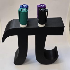 Puede incluir: Un organizador de escritorio negro con forma del símbolo matemático pi, que contiene cuatro bolígrafos de colores. Los bolígrafos son turquesa, azul, morado y negro, cada uno con una base blanca y una tapa a juego.