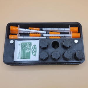 Puede incluir: Un organizador de plástico negro que contiene varias jeringas con tapas naranjas, una almohadilla con alcohol y varios objetos hexagonales de color gris oscuro. Las jeringas son transparentes con marcas negras y naranjas.