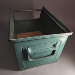 Op de afbeelding: Een rechthoekige opbergbak van blauwgroen metaal met een verzonken handgreep op het voorpaneel, een bovenrand met een horizontale stang en een platte bodem. Het metalen oppervlak vertoont tekenen van slijtage en oxidatie met verfschilfers.