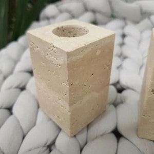 Peut inclure: Un bougeoir carré en pierre beige avec une ouverture circulaire sur le dessus. La pierre a une texture poreuse et un aspect en couches. Le bougeoir est présenté sur une surface blanche tressée.
