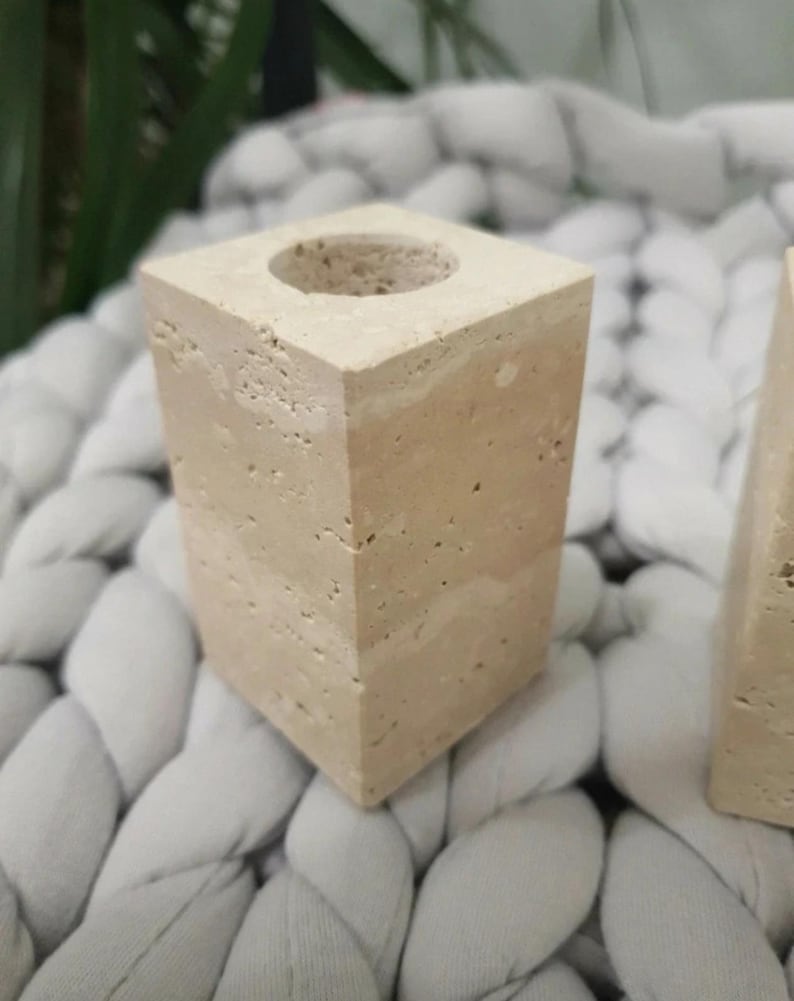 May include: A square, beige stone candle holder with a circular opening at the top. The stone has a porous texture and a layered appearance. The candle holder is displayed on a white, braided surface.