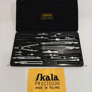 May include: A black rectangular case containing a set of silver metal drafting instruments, including various compasses and dividers. The interior lid features a gold label reading Skala PRECISION MADE IN POLAND, matching the yellow cloth below.