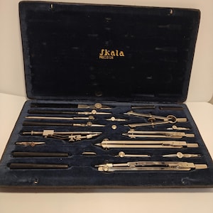 Può includere: Un set da disegno vintage in una custodia foderata in velluto blu scuro. Il set include vari compassi, divisori e altri strumenti da disegno color argento. La custodia è contrassegnata con "skala PRECISION" in lettere dorate.