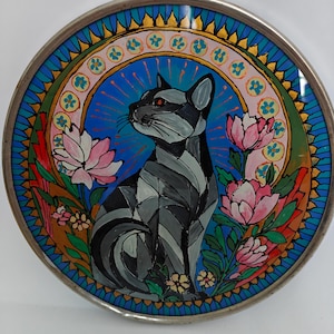Può includere: Un piatto decorativo circolare con un'illustrazione in stile vetrata raffigurante un gatto grigio e nero seduto al centro. Il gatto è circondato da fiori di loto rosa, foglie verdi e uno sfondo blu con un bordo floreale giallo e rosa.