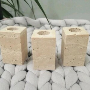 May include: Three beige stone candle holders with a square shape and a circular opening at the top. The candle holders are displayed on a white, textured surface.
