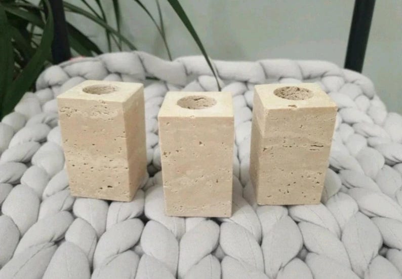 May include: Three beige stone candle holders with a square shape and a circular opening at the top. The candle holders are displayed on a white, textured surface.