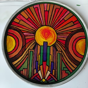 Könnte beinhalten: Ein rundes Dekotablett mit einem handgemalten Motiv einer stilisierten Sonne, die über einer geometrischen Stadtlandschaft aufgeht. Das Kunstwerk verwendet leuchtende Rot-, Orange-, Gelb-, Grün- und Violetttöne in einem silbernen Rahmen.