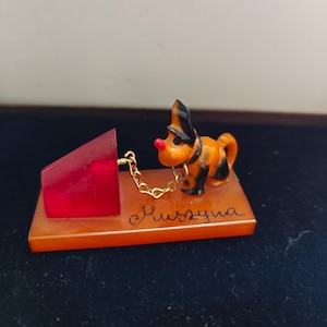 Può includere: Statuetta vintage raffigurante un piccolo cane arancione e nero con un naso rosso e una catena d'oro, attaccato a un blocco rosso. Il cane è su una base rettangolare arancione con testo corsivo.