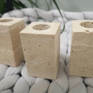 May include: Three beige stone candle holders with a square base and a circular opening at the top. The candle holders are made of a porous stone material and are arranged in a row. The background is a light gray, textured surface.