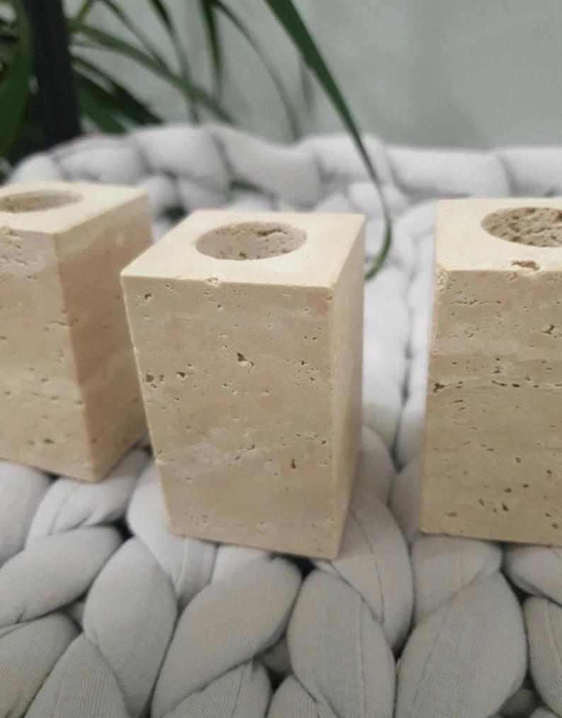 May include: Three beige stone candle holders with a square base and a circular opening at the top. The candle holders are made of a porous stone material and are arranged in a row. The background is a light gray, textured surface.