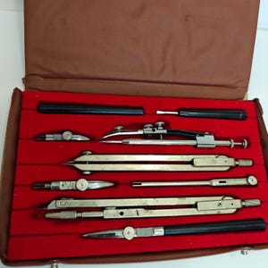 Könnte beinhalten: Ein Vintage-Zeichensatz in einem braunen Koffer mit rotem Samtinterieur. Das Set enthält mehrere silberfarbene und schwarze Zirkel und Teiler in verschiedenen Größen, die ordentlich in ihren vorgesehenen Schlitzen angeordnet sind. Der Koffer ist geöffnet und zeigt die Werkzeuge.
