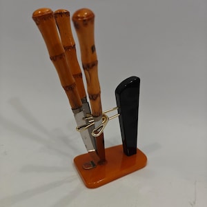 Op de afbeelding: Een set van drie messen met handvatten in bamboestijl en roestvrijstalen lemmeten. De messen worden gepresenteerd in een standaard met een zwarte staander en een oranje basis. De standaard heeft een goudkleurige metalen houder voor de messen.