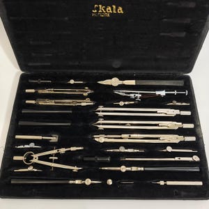 Può includere: Un set da disegno vintage in una custodia foderata in velluto nero. Il set include vari strumenti da disegno argentati e neri, come compassi, divisori e penne. La custodia è aperta, rivelando gli strumenti.
