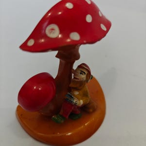 Op de afbeelding: Een grillig beeldje met een rode paddenstoel met witte stippen en een kleine rode paddenstoel. Een lachende kabouterfiguur zit onder de paddenstoel. De basis is oranje.