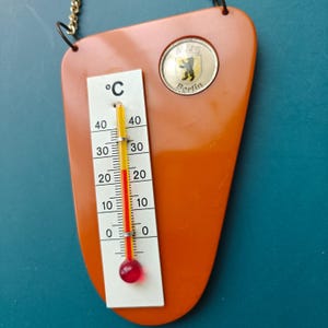 Puede incluir: Un termómetro de pared naranja de forma triangular con una escala blanca marcada en grados Celsius. El dispositivo cuenta con un tubo de vidrio central con líquido rojo y un emblema circular en la parte superior con un oso y el texto Berlin.