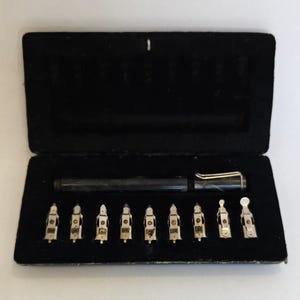 May include: A black pen and several silver nibs are displayed inside a black velvet case. The pen has a silver clip and a black body. The nibs are small and have a unique shape, likely for calligraphy or drawing.