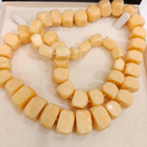 May include: A necklace composed of light peach-colored, rectangular beads. The beads are strung together, forming a double strand. The necklace is displayed in a white box, showcasing its color and shape.
