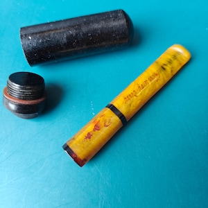 Puede incluir: Una pluma estilográfica jaspeada en amarillo y negro con un capuchón negro separado y una pequeña pieza final roscada de color negro. El cuerpo de la pluma presenta el texto en cursiva "Swan Ivory Carry" en un tono oscuro sobre el fondo amarillo.
