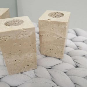 May include: Two beige stone candle holders with a square base and a circular indentation at the top. The candle holders are made of a textured stone material and are displayed on a light gray, woven surface.