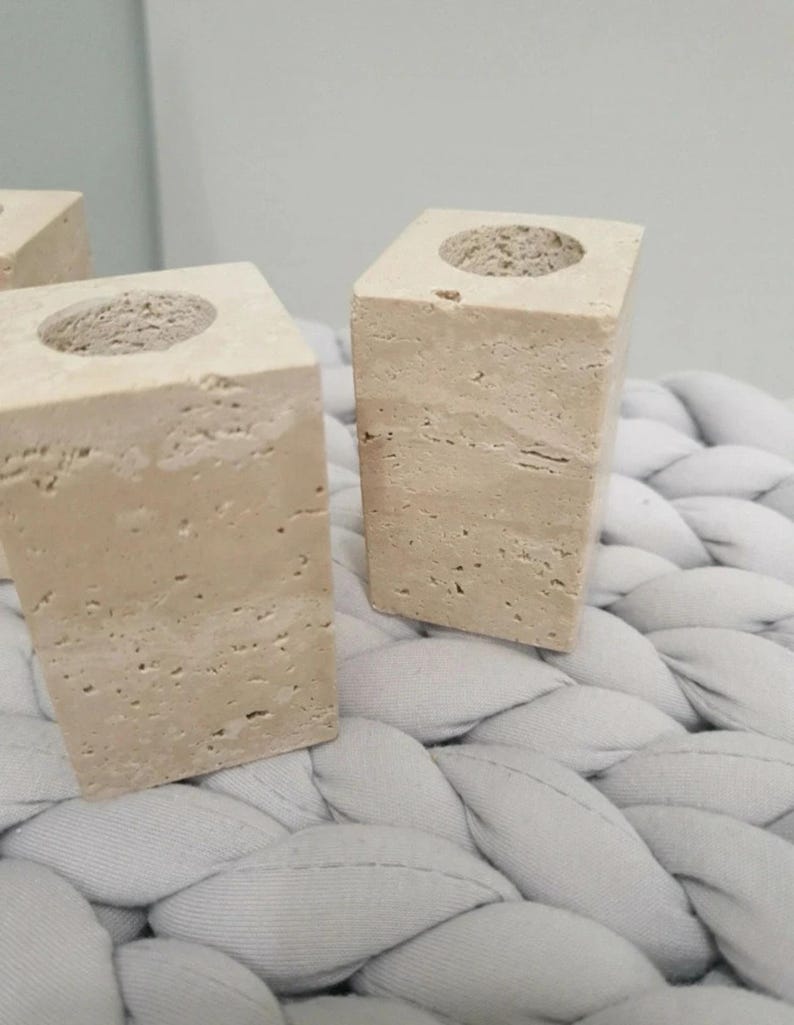 May include: Two beige stone candle holders with a square base and a circular indentation at the top. The candle holders are made of a textured stone material and are displayed on a light gray, woven surface.