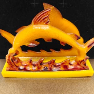 Könnte beinhalten: Ein gelber Serviettenhalter mit Delfin-Motiv. Zwei Delfine springen über Wellen, mit gelbem Sockel und bernsteinfarbenen Akzenten. Der Serviettenhalter besteht aus einem durchscheinenden Material.