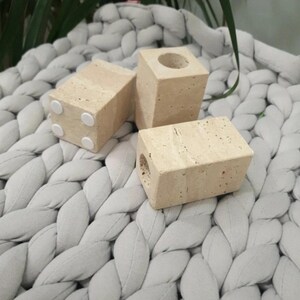 May include: Three beige, rectangular candle holders with a porous texture. One has a circular opening at the top, and another has a hole on the side. The candle holders have small, white, circular pads on the bottom. The objects are on a gray, braided textile.