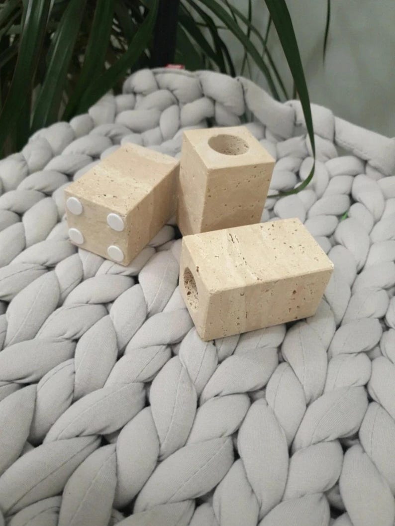 May include: Three beige, rectangular candle holders with a porous texture. One has a circular opening at the top, and another has a hole on the side. The candle holders have small, white, circular pads on the bottom. The objects are on a gray, braided textile.