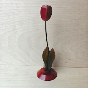 Könnte beinhalten: Eine dekorative Tulpen-Skulptur mit einem glänzend roten, zwiebelförmigen Kopf, einem dünnen schwarzen Stiel und zwei dunkelbraunen, durchscheinenden Blättern, die aus einem runden roten Sockel ragen. Das Objekt hat glatte, polierte Oberflächen.